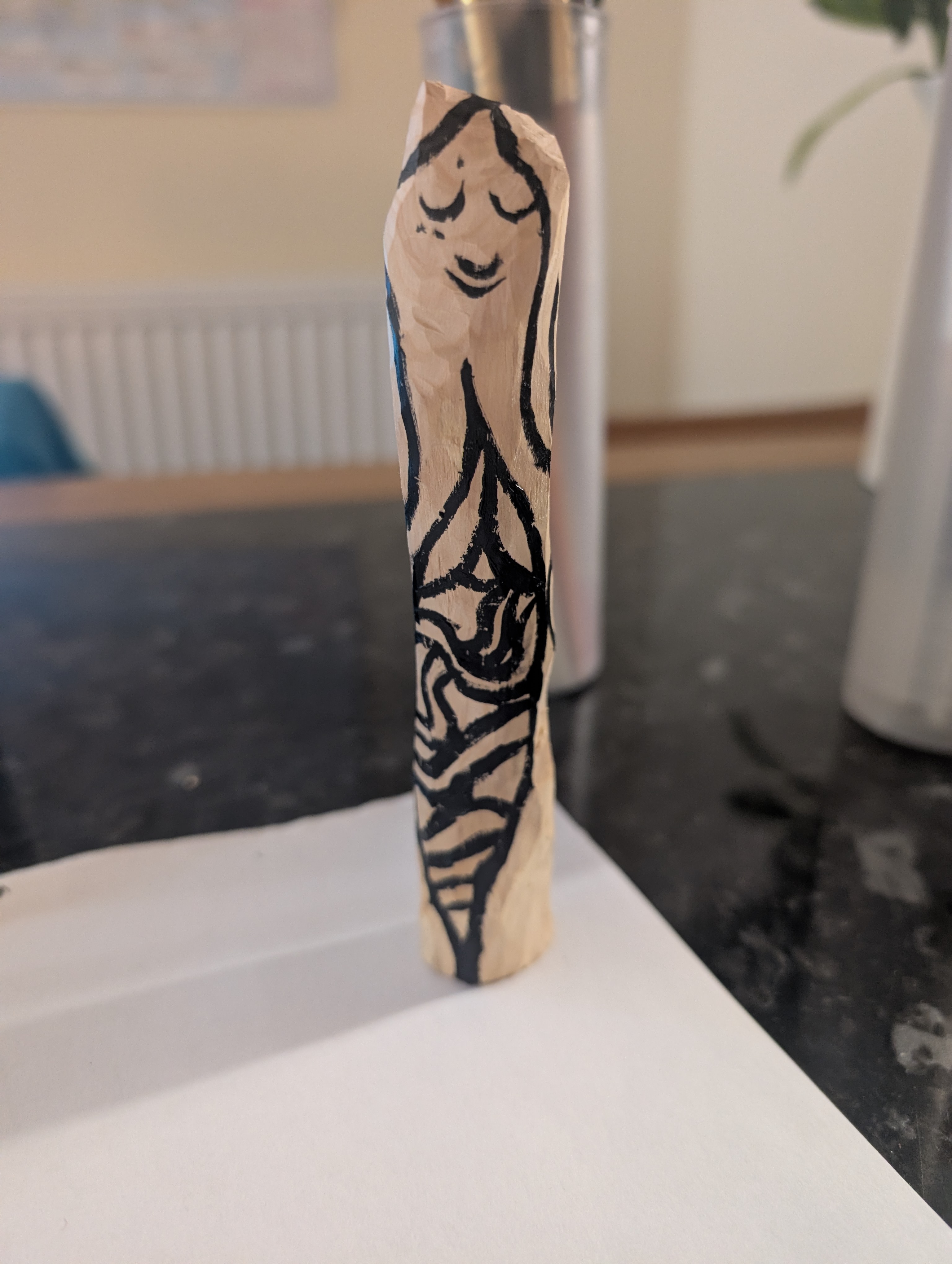 A wooden figure of a young man with a rested face and closed eyes, he is made from a phallic shaped piece of carved wood and has been painted on with black oil paint